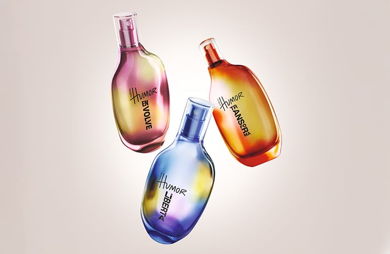 Foto de tres frascos de perfume Humor de Natura, en tonos vibrantes de rosa, naranja y azul, con diseño irregular, sobre un fondo beige claro.